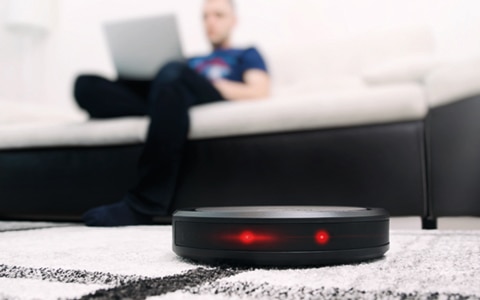 Automatic vaccum cleaner in foreground with blurred image of man sitting on sofa cross legged