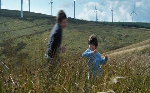 two boys running through long grass with wind turbines in background